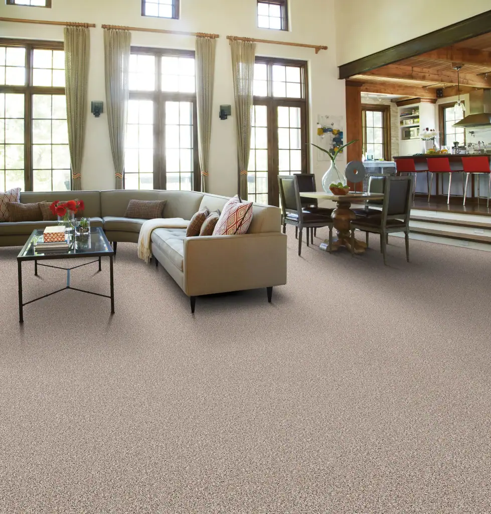 A spacious living room with large windows, beige carpet by Flooring Installation Wake County, a light beige sectional sofa, a glass coffee table, decorative pillows, and an open dining area with modern chairs in the background.